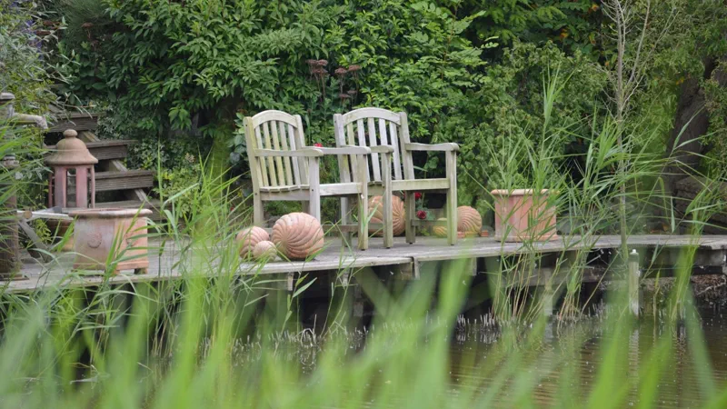 Holzsteg mit zwei Gartenstühlen am Teich, davor Schilf