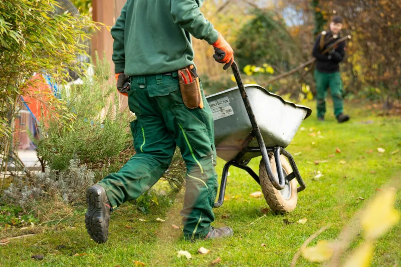 Landschaftsgärtner in grüner Arbeitskleidung zieht eine Schubkarre durch einen Garten, ein Kollege arbeitet im Hintergrund
